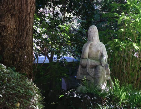 A stone statue of a seated figure surrounded by lush greenery and trees in a serene outdoor setting.