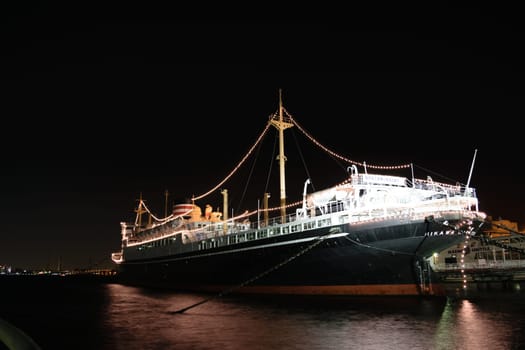 A large, illuminated ship docked at a pier at night. The ship is adorned with string lights, creating a festive atmosphere. The water reflects the lights, adding to the scenic view.