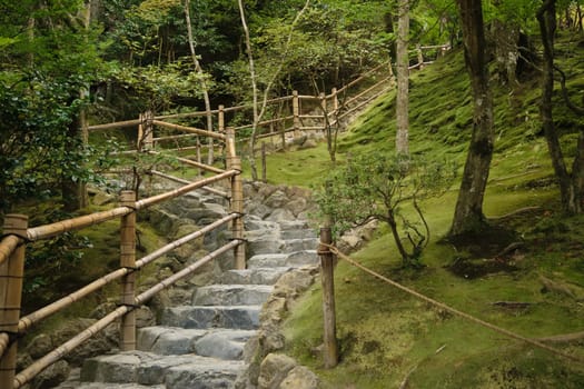 A stone pathway with wooden railings winding through a lush, green forest area. The path is surrounded by moss-covered ground and various trees.