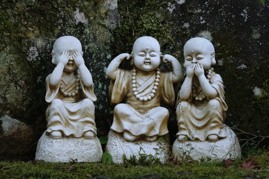 Three small stone statues of monks in a garden, each representing the 'see no evil, hear no evil, speak no evil' proverb. The statues are placed against a mossy rock background.