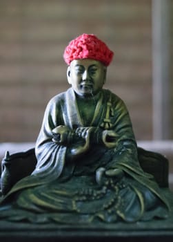 A statue of a seated monk wearing a red knitted hat. The monk is holding an object in his hands and appears to be in a meditative pose. The background is blurred.