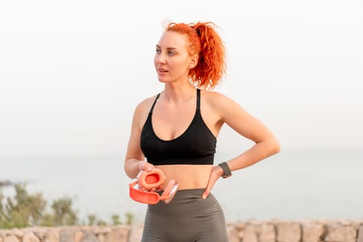 Sporty woman with red hair holding wireless headphones taking a break after tiring morning jogging. Fit female jogger in sportswear looking away. Health and fitness concepts.