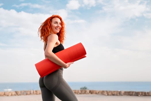 Smiling redhead sportswoman holding rolled yoga mat. Side view of fit strong female athlete in sportswear looking away with sea in background under cloudy sky on sunny day.