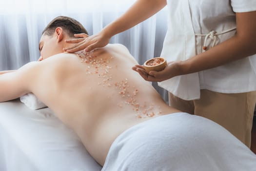 Blissful man customer having exfoliation treatment in luxury spa salon with warmth candle light ambient. Salt scrub beauty treatment in health spa body scrub. Quiescent