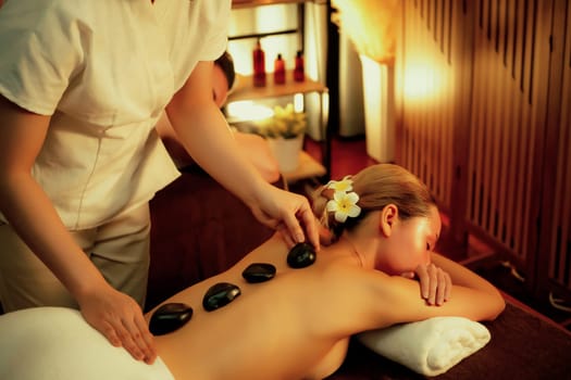 Hot stone massage at spa salon in luxury resort with warm candle light, blissful woman customer enjoying spa basalt stone massage glide over body with soothing warmth. Quiescent