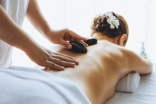 Hot stone massage at spa salon in luxury resort with day light serenity ambient, blissful woman customer enjoying spa basalt stone massage glide over body with soothing warmth. Quiescent