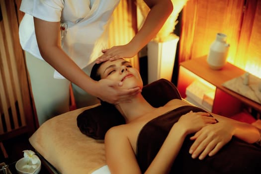 Caucasian woman enjoying relaxing anti-stress head massage and pampering facial beauty skin recreation leisure in warm candle lighting ambient salon spa in luxury resort or hotel. Quiescent