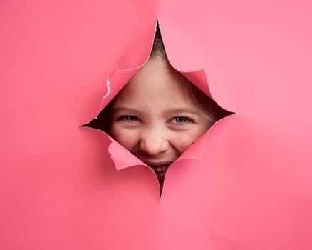 Cute Caucasian girl peeks out of a hole in a paper pink background