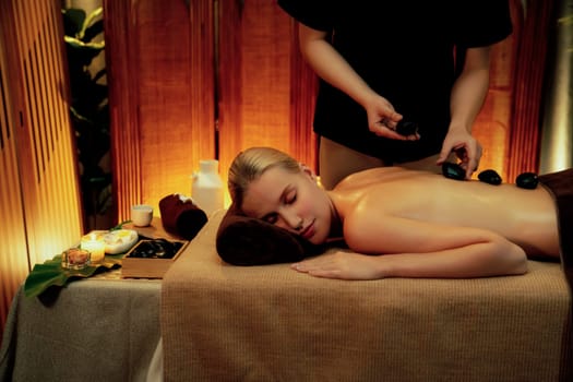 Hot stone massage at spa salon in luxury resort with warm candle light, blissful woman customer enjoying spa basalt stone massage glide over body with soothing warmth. Quiescent