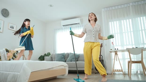 Caucasian mom and asian child doing housework together and dancing to music. Attractive mother and daughter cleaning modern bedroom while sing along and jump to relaxing music. Lifestyle. Pedagogy.