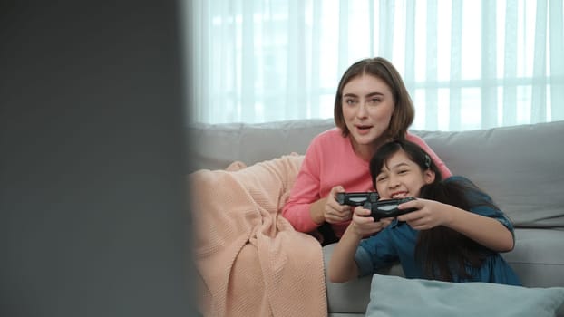 Happy girl playing game and sitting while mother lie on sofa. Caucasian parent and attractive smart girl spending time together while holding joystick and focus on winning games. Lifestyle. Pedagogy.