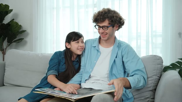 Happy daughter lean on father shoulder while listening story telling. Young asian schoolgirl looking ar dad while reading story book together and sitting at sofa. Family recreation concept. Pedagogy.