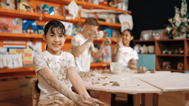 Cute girl smiling and sitting while diverse friend doing pottery workshop. Highschool student wearing dirty shirt while looking at camera at workshop in art lesson. Blurring background. Edification.