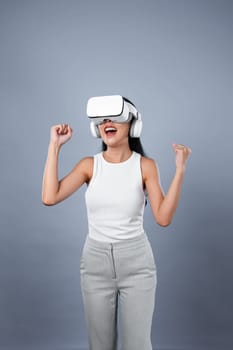 Smart female standing with gray background wearing VR headset connecting metaverse, futuristic cyberspace community technology. Elegant woman excited seeing generated virtual scenery. Hallucination.