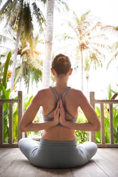 Back view of young woman doing reverse namaste mudra while meditating in tropical vacation resort. Reverse prayer pose. Vertical image. Yoga and spirituality concepts.