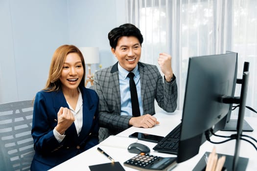 Successful smiling Asian business partners raising fist up together in profitable project job in stock exchange database on pc wearing formal suit at modern home office at side view image. Infobahn.