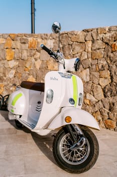 White vintage motor scooter stands against a stone wall on the sidewalk. High quality photo