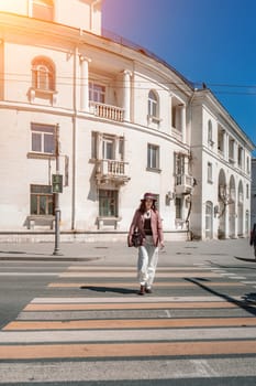 Woman city road crossing. Stylish woman in a hat crosses the road at a pedestrian crossing in the city. Dressed in white trousers and a jacket with a bag in her hands