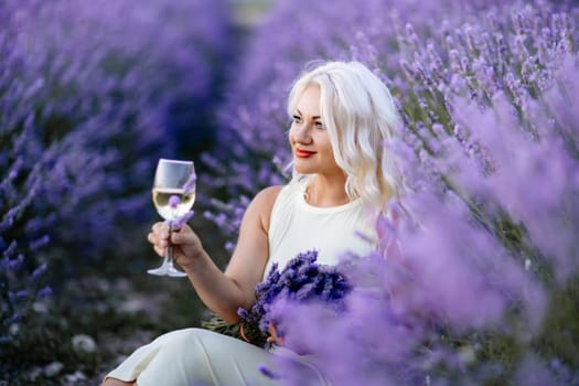 Blonde lavender field holds a glass of white wine in her hands. Happy woman in white dress enjoys lavender field picnic holding a large bouquet of lavender in her hands . Illustrating woman's picnic in a lavender field