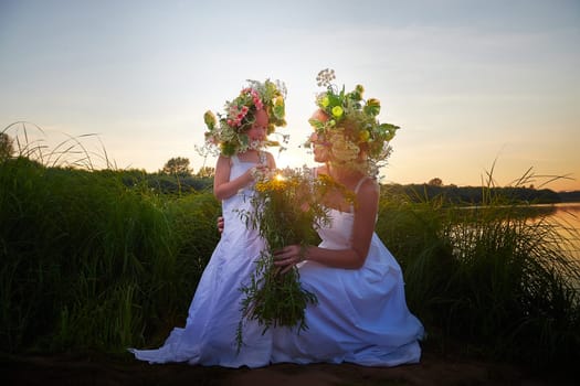 Mother and daughter in white sundresses, wreaths of flowers and bouquet in nature evening at sunset . Family celebrate Slavic Holiday of Ivan Kupala