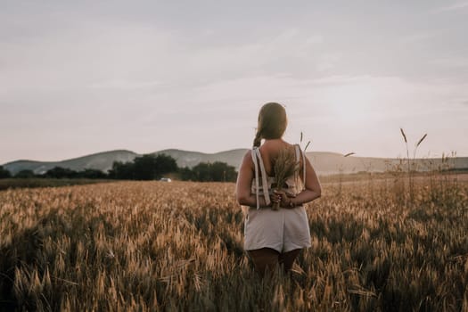 A woman is holding a bunch of wheat in her arms. The wheat is dry and brown, and the woman is wearing a white dress. The scene is set in a field, and the woman is posing for a photo