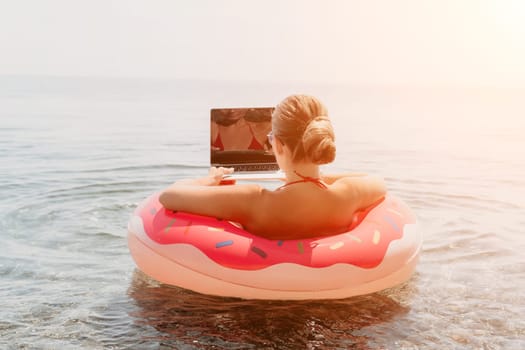 Woman freelancer works on laptop swimming in sea on pink inflatable ring. Pretty lady typing on computer while floating in the sea on inflatable donut at sunset. Freelance, remote work on vacation