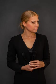 A woman in a black dress holding a pair of makeup brushes. She is looking at the camera