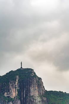 Christ the Redeemer stands atop a cliff; tourists flock at its base under a cloudy sky.