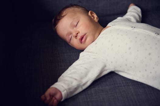 Close-up authentic portrait of a cute baby boy in white bodysuit, sweetly sleeping on comfortable gray sofa. The concept of happy childhood. Children. Infancy and babyhood. World Children's Day