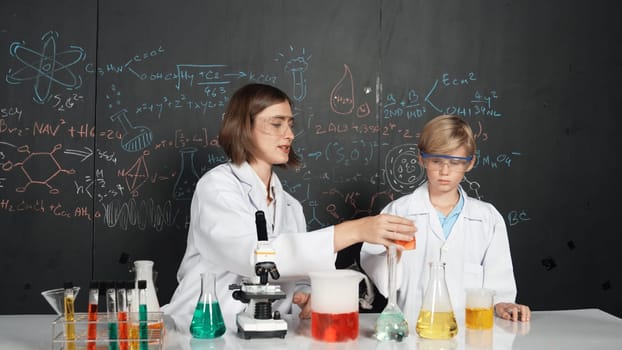 Caucasian teacher helps student mixed liquid in science laboratory. Professional instructor support smart boy doing experiment in STEM science class at blackboard written chemical theory. Erudition.