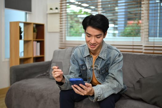 Smiling young Asian man doing online payment through credit card and smart phone.