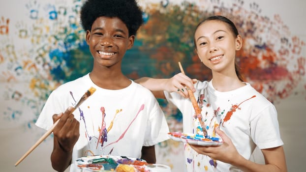 Highschool student with mixed races standing and looking at camera at art lesson. Diverse children hold the paintbrush while pose with confident at colorful stained wall. Creative style. Edification.