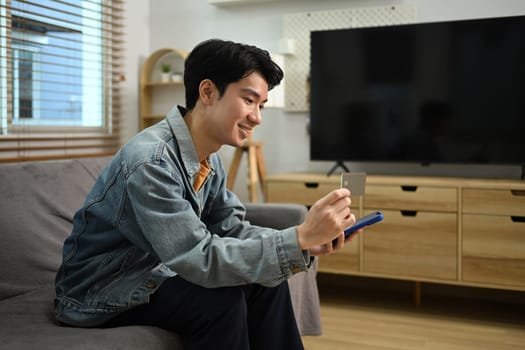 Smiling young Asian man with credit card making financial transaction on smart phone.