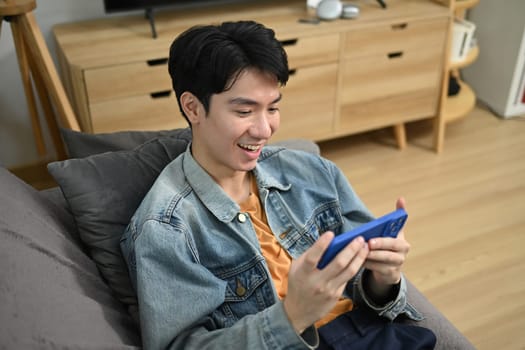 Smiling man playing video game on smart phone while sitting on couch at home.