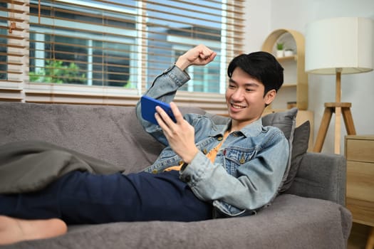 Happy young man looking at smartphone and celebrating good news sitting on couch at home.