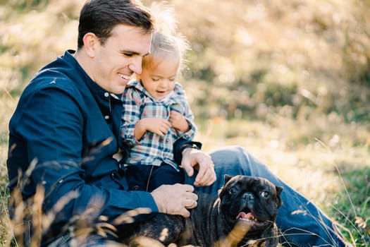 Little girl sitting on dad lap petting french bulldog. High quality photo