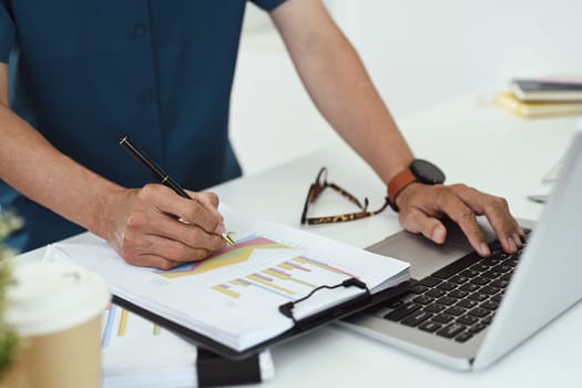 Cropped shot professional businessman analyzing graph paper and using laptop at desk.