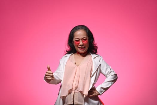 Confident cool senior woman in trendy glasses showing thumbs up gesture over pink background.