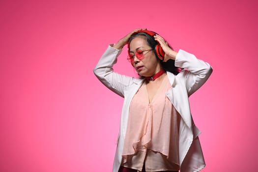 Happy stylish middle age woman listening to music with wireless headphones over pink background.