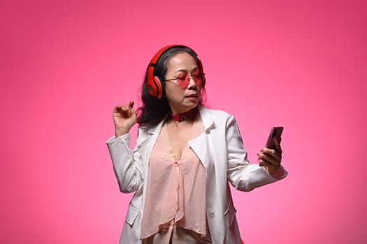 Carefree senior woman listening to music in headphones using mobile phone on pink background.