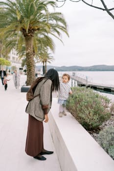 Mom leaned over to the little girl standing on the border of a green flower bed on the boardwalk. Back view. High quality photo