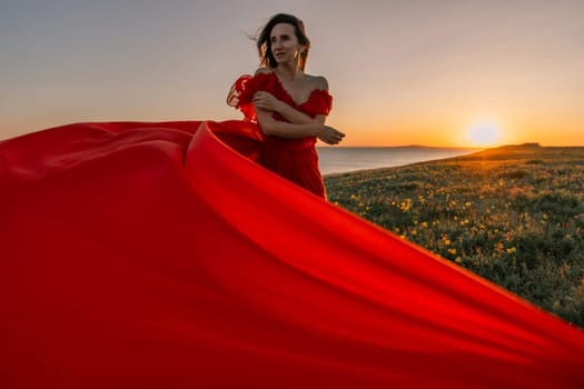 woman red dress standing grassy hillside. The sun is setting in the background, casting a warm glow over the scene. The woman is enjoying the beautiful view and the peaceful atmosphere