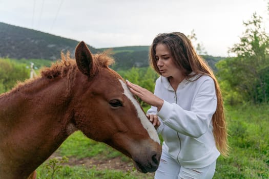 A woman is petting a brown horse. The woman is wearing a white jacket. The horse is standing in a grassy field