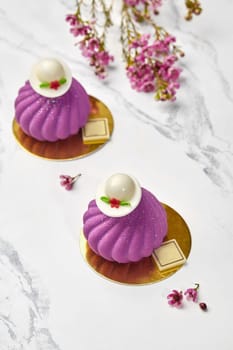 Elegant purple mousse desserts topped with white chocolate hats arranged on golden cardboards, accompanied by delicate spring blossoms on marble surface