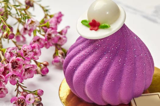 Closeup of tempting swirl-shaped mousse pastry with velvety texture topped with elegant white chocolate hat, presented on golden cardboard, accompanied by sprig of delicate violet flowers