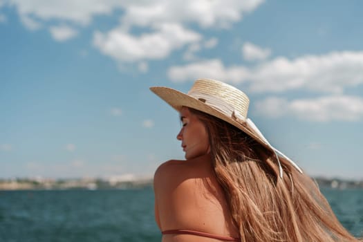 A woman in a swimsuit sits with her back holding a hat, looks at the ocean, sunny day, relaxes