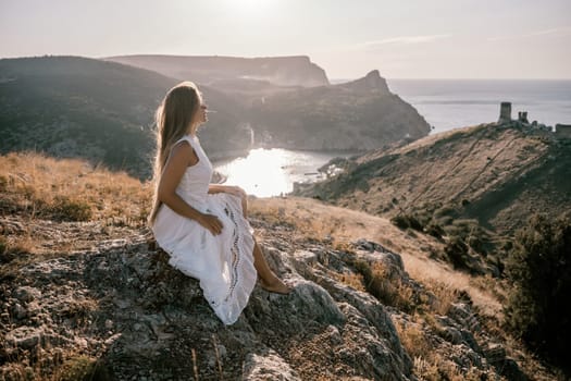 A woman in a white dress sits on a rock overlooking a body of water. The scene is serene and peaceful, with the woman enjoying the view and the calmness of the surroundings