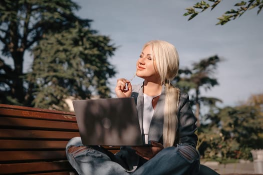 A blonde woman sits on a bench with a laptop in front of her. She is wearing a gray jacket and jeans. The scene suggests a casual and relaxed atmosphere, as the woman is using her laptop outdoors