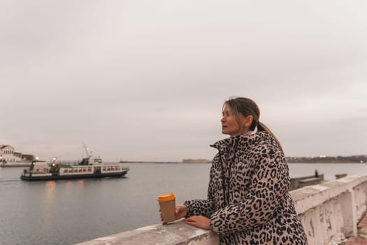 A woman in a leopard print coat stands on a ledge overlooking a body of water. She is holding a cup of coffee and looking out at the water. The scene is peaceful and serene