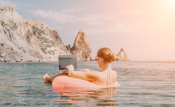 Woman freelancer works on laptop swimming in sea on pink inflatable ring. Pretty lady typing on computer while floating in the sea on inflatable donut at sunset. Freelance, remote work on vacation